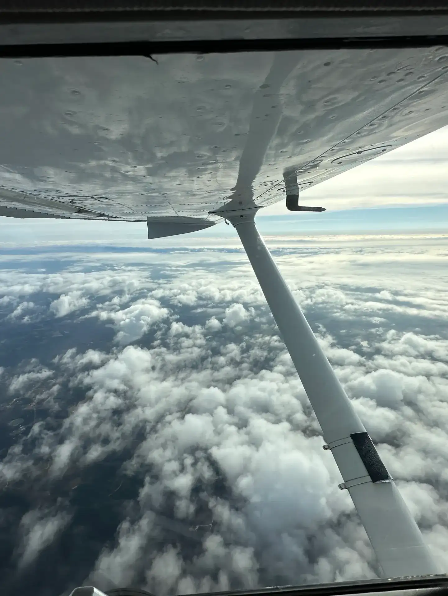 View from inside a training airplane of a wing and strut above scattered clouds