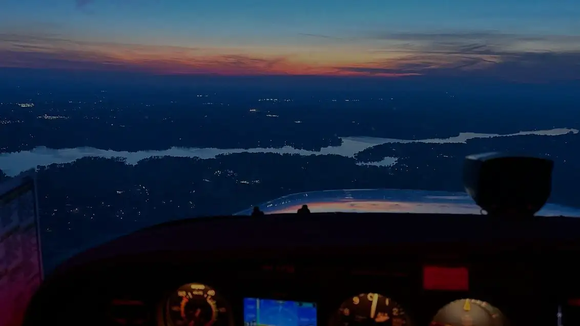 Dusk view from a training aircraft cockpit over a lake and dark wooded land with an orange sunset on the horizon